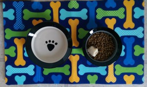 Ceramic food and water bowls for the dog, on a mat decorated with brightly colored bones.