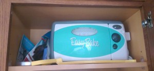 Blue and white plastic Easy-Bake toy oven on a kitchen cabinet shelf, with its accessories.