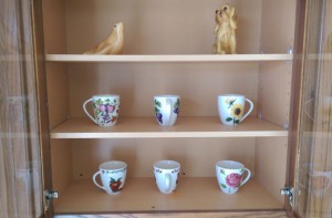 Kitchen cabinet with three shelves and glass doors, open to show brightly painted mugs and candle-wax animal figurines.