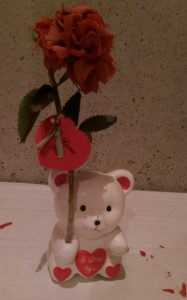 Ceramic bear with "I Love You" on its chest, holding a decayed fake flower.