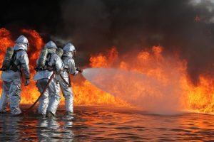 Three firefighters do a training exercise with flames in background.