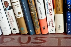 Photo of books on a shelf with the word "DUST" written in the dust.