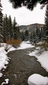 Photo of river with snowy banks, conifers, and a mountain range.